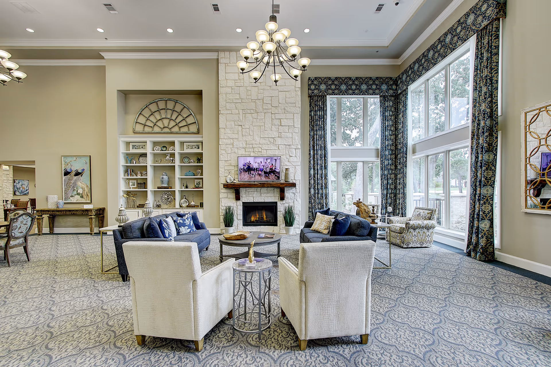 A spacious and elegant living room area with large windows allowing natural light to fill the space. The room features two white armchairs and two dark blue sofas arranged around a round coffee table in front of a stone fireplace with a mounted TV above it. There are decorative shelves with various ornaments, patterned curtains, and a chandelier hanging from the ceiling. The carpet has a detailed pattern, and there is additional seating and artwork visible in the background.