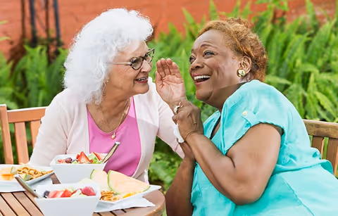 Two elderly women sitting at an outdoor wooden table, smiling and enjoying a meal together with bowls of fruit and snacks in front of them, surrounded by green plants.