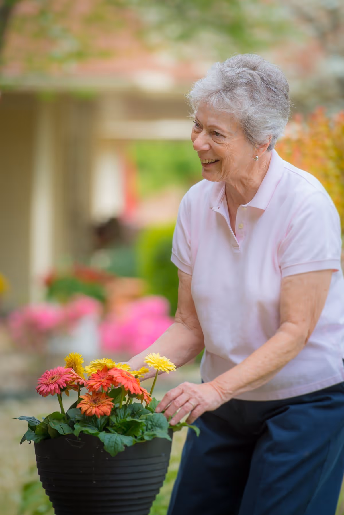 An elderly woman with short gray hair wearing a light pink polo shirt and dark pants is smiling while tending to a black pot filled with colorful flowers including pink, orange, and yellow blooms in an outdoor garden setting.