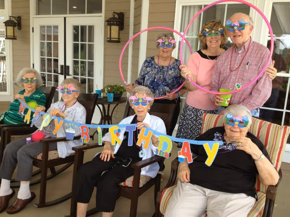 A group of six elderly people and one middle-aged woman celebrating a birthday on a covered patio. They are all wearing colorful, festive glasses with flamingo and star designs. Four elderly women are seated in chairs holding a colorful 'HAPPY BIRTHDAY' banner, while one elderly woman and the middle-aged woman stand behind them holding pink hula hoops. The elderly man standing next to the middle-aged woman is holding a green cup. The setting appears to be at Oak Ridge Senior Living Community.