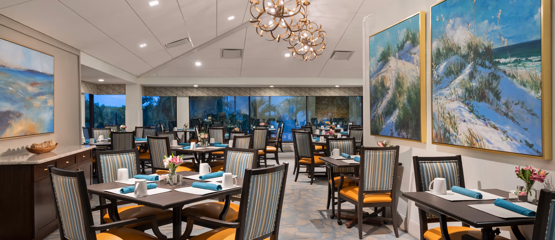 A spacious dining room with multiple tables set with white mugs, blue napkins, and small flower arrangements. The chairs have striped upholstery with orange cushions. Large windows line the back wall, and colorful paintings of beach scenes hang on the walls. The ceiling has modern light fixtures.