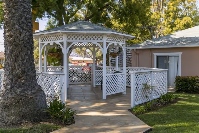 White wooden gazebo with lattice panels situated on a tiled pathway surrounded by green grass, plants, and trees, adjacent to a beige building with a sliding glass door.