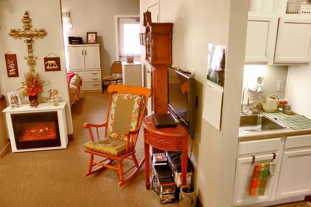 Cozy senior living area with a rocking chair, TV on a small table, an electric fireplace, and a kitchenette visible on the right.