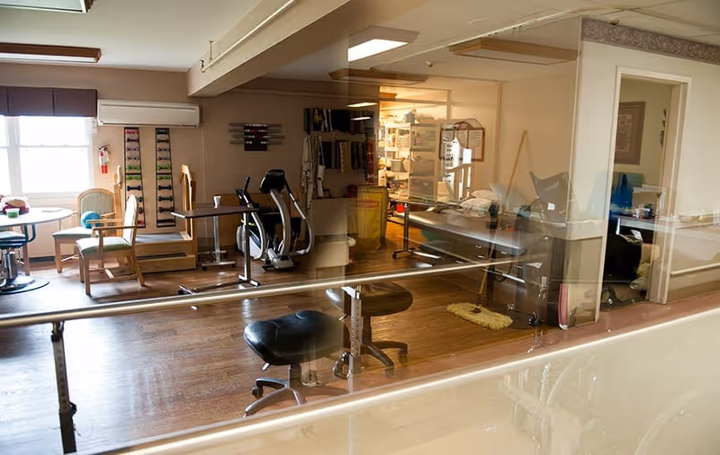 Interior view of a rehabilitation and skilled nursing center room with exercise equipment, chairs, tables, and various therapy tools. The room has wooden flooring and is well-lit by natural light from a window. There is a glass partition in the foreground and an adjacent room visible through an open doorway.