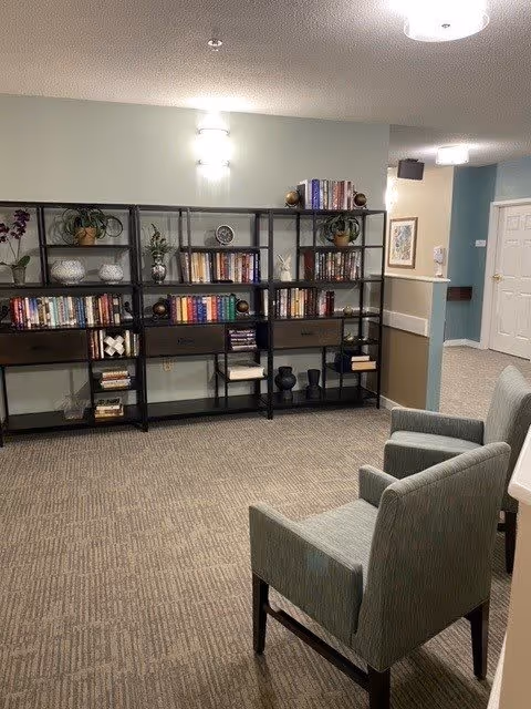 A quiet interior room with two green upholstered chairs facing a black metal bookshelf filled with books, decorative plants, and ornaments. The room has beige carpet, light-colored walls with a chair rail, and ceiling lights. A hallway with a white door and framed artwork is visible in the background.