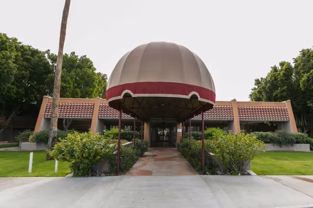 Front exterior view of Rio Vista Post Acute And Rehabilitation building with a large beige and maroon canopy over the entrance walkway, surrounded by green bushes and trees.