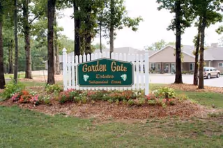 A white picket fence sign with green background and yellow text reading 'Garden Gate Estates Independent Living' surrounded by flowers and trees, with a building and parked cars visible in the background.