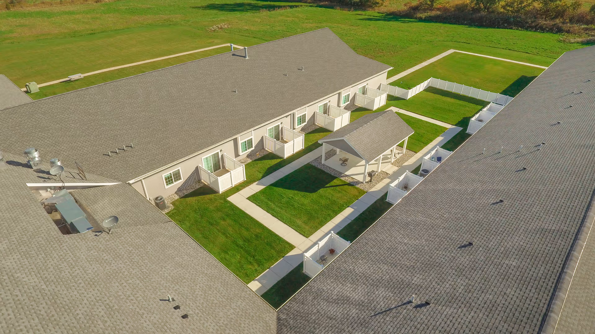 Aerial view of a senior living facility courtyard with two long buildings facing each other. The courtyard features green grass, paved walkways, a covered gazebo with seating, and small fenced patio areas attached to each unit.