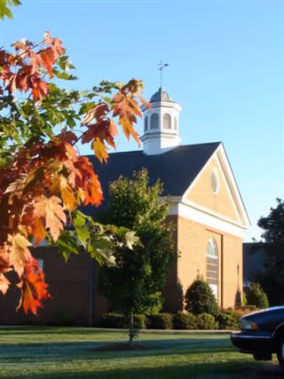 Exterior view of a brick building with a white cupola on the roof, surrounded by trees with green and autumn-colored leaves, under a clear blue sky. Part of a car is visible in the lower right corner.