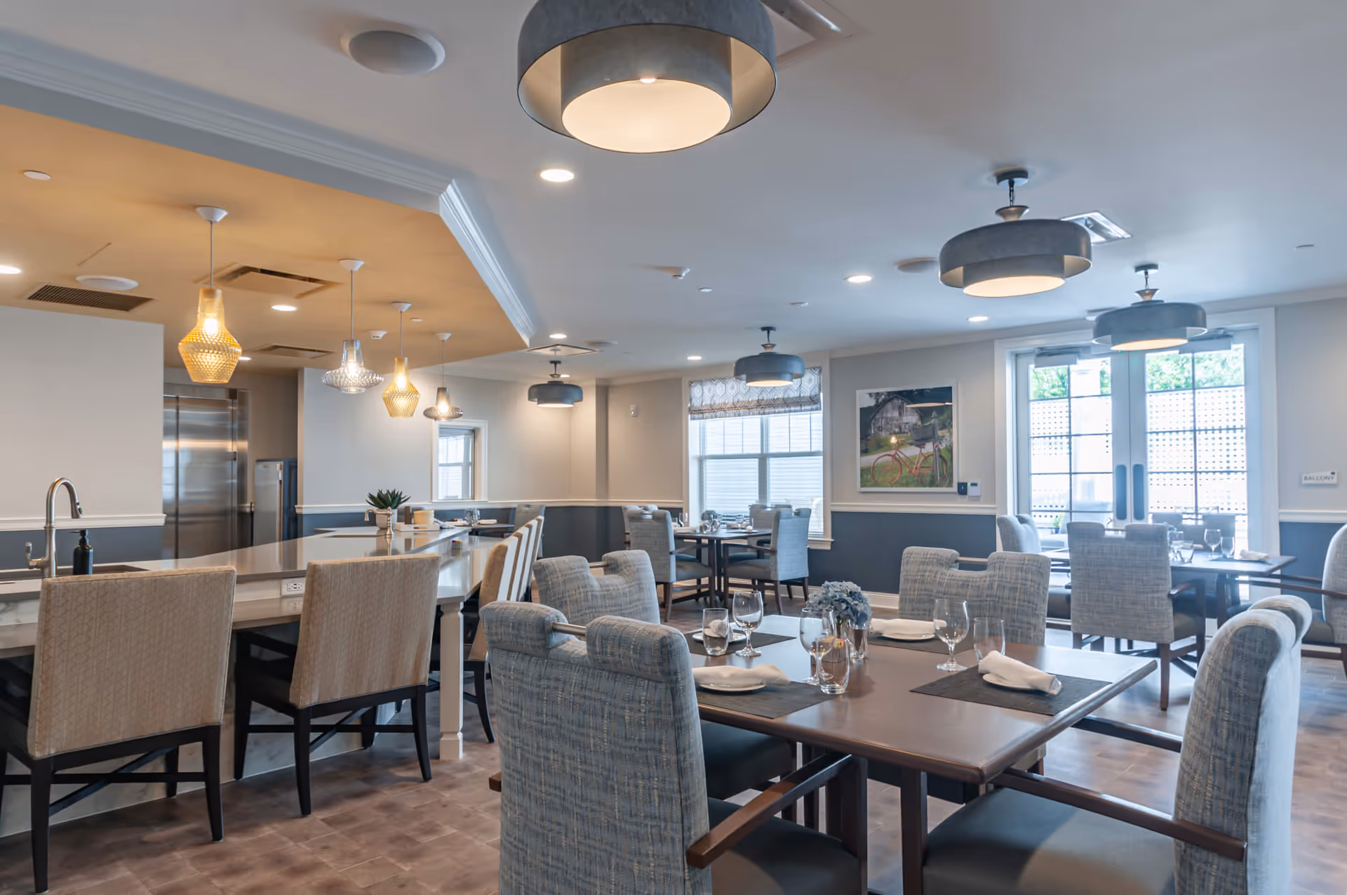 A modern dining room with multiple tables and chairs arranged neatly. The tables are set with placemats, napkins, and glassware. There is a kitchen island with high chairs on the left side, pendant lights hanging above it, and large windows and glass doors letting in natural light. The walls are painted in a two-tone color scheme with artwork and a sign indicating a balcony.