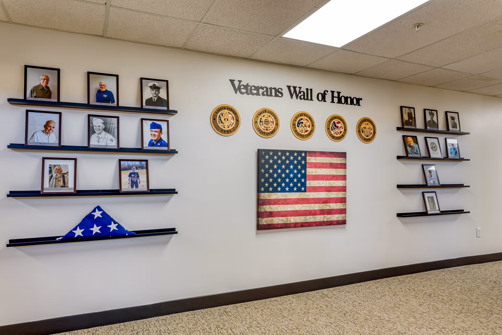 A hallway wall display titled 'Veterans Wall of Honor' featuring framed photographs of veterans, military insignias for different branches of the United States Armed Forces, and a folded American flag on shelves beneath the photos.