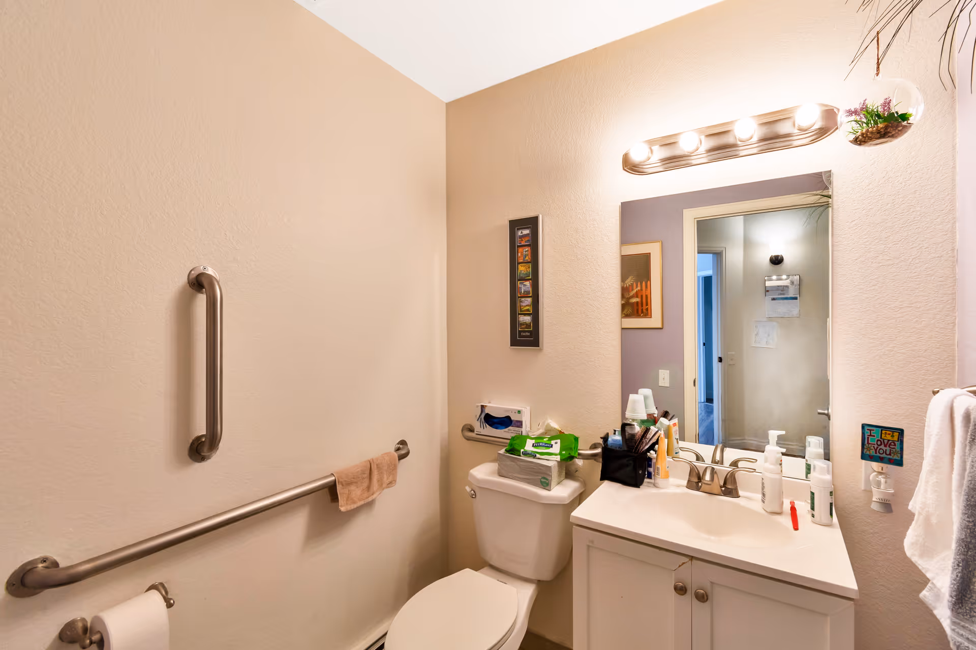 Small accessible bathroom with a toilet, sink vanity, mirror, grab bars and toiletries on the counter.