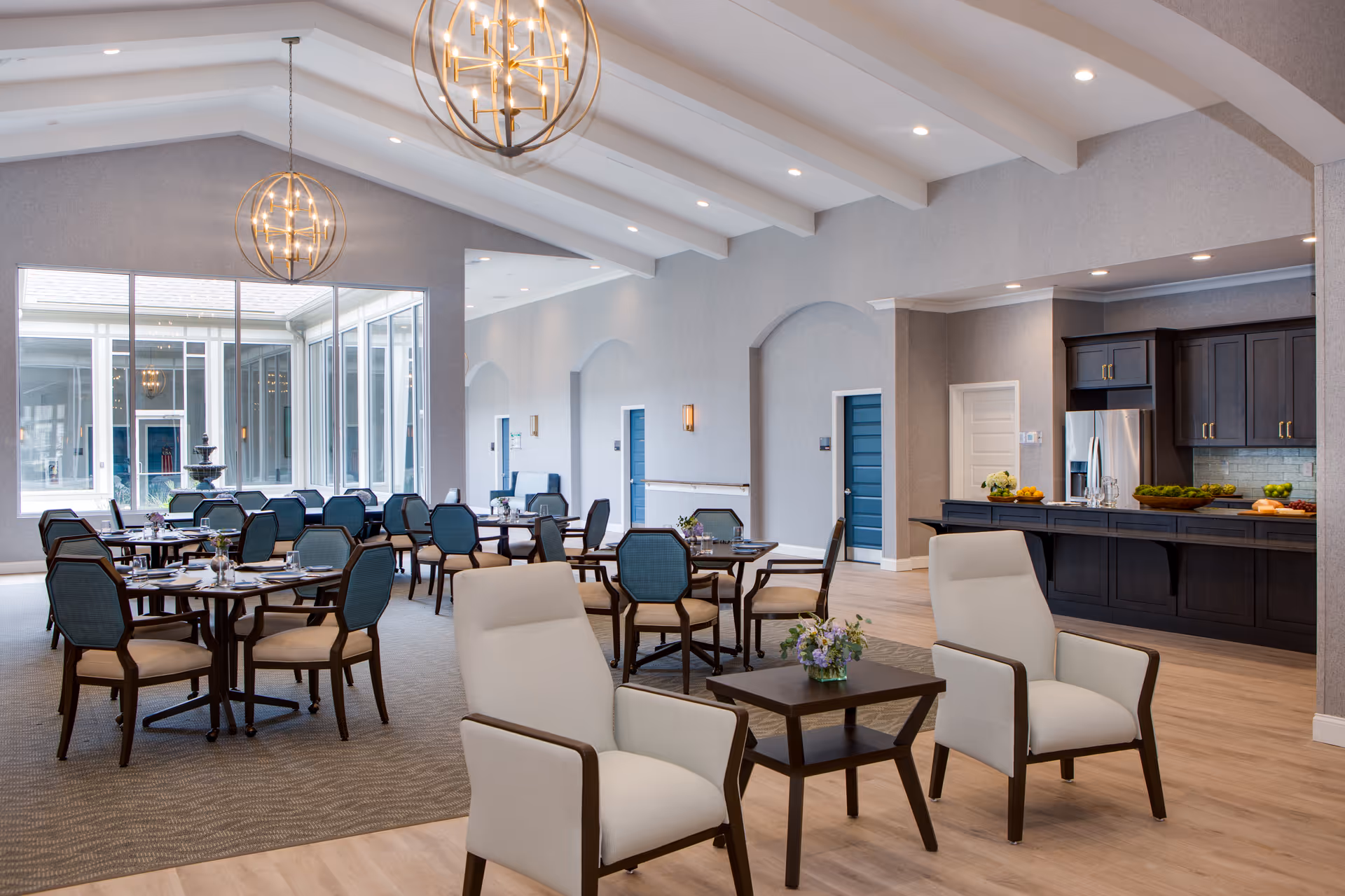 A spacious and well-lit dining area in a senior living facility featuring multiple round tables with blue cushioned chairs, two beige armchairs with a small wooden table between them, modern spherical pendant lights hanging from a vaulted ceiling, large windows allowing natural light, and a kitchen area with dark cabinetry and stainless steel appliances in the background.