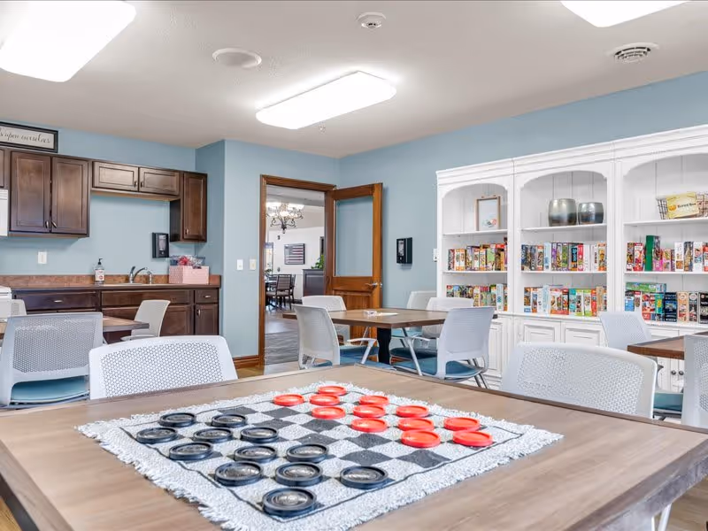 A bright and spacious common area with light blue walls, white ceiling lights, and wooden trim. The room features a wooden table in the foreground with a checkers game set up, surrounded by white chairs. In the background, there is a kitchenette with dark wood cabinets and a sink, as well as a white shelving unit filled with board games. An open wooden door leads to another room with a dining table and chairs.