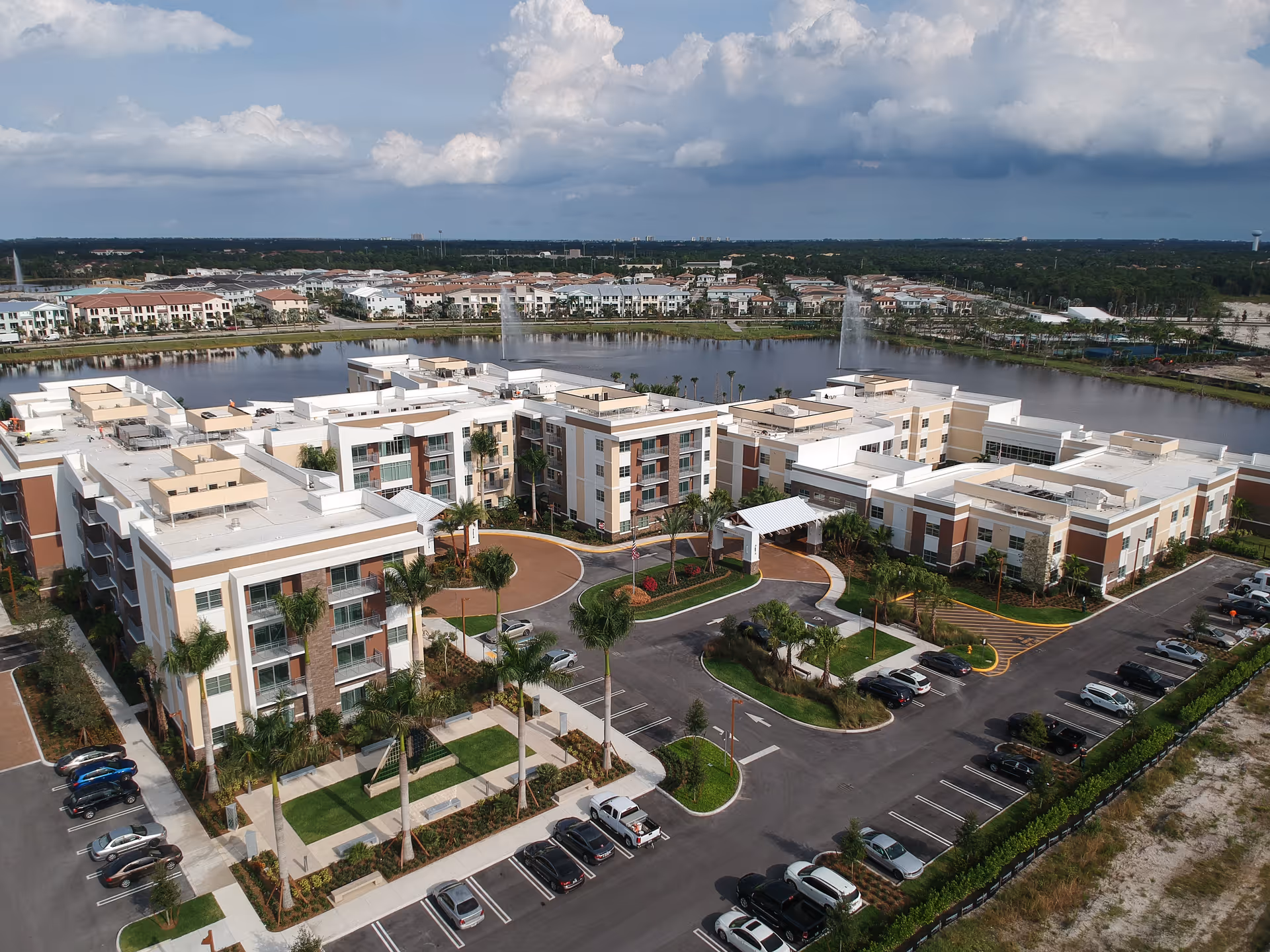 Aerial view of a modern multi-story senior living complex by a lake with fountains, landscaped courtyards, and surrounding parking.