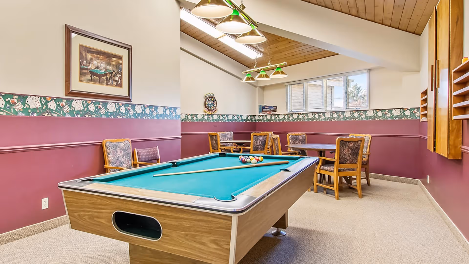 A recreational room with a pool table in the center, surrounded by chairs and tables. The walls are painted with a maroon lower half and beige upper half, decorated with a patterned wallpaper border featuring playing cards and dice. The ceiling is wooden with hanging green and white lights above the pool table. A framed picture hangs on the left wall.