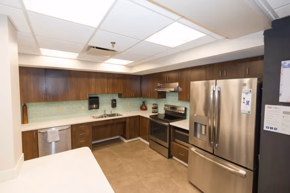 A modern kitchen with dark wood cabinets, light countertops, a stainless steel refrigerator, stove, and dishwasher. The backsplash is made of light green subway tiles, and there is a double sink under the cabinets. The ceiling has recessed lighting panels.