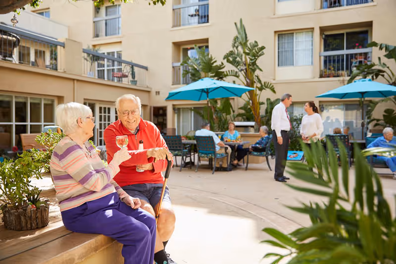 Two elderly people sitting on a bench in an outdoor courtyard of a senior living facility, enjoying drinks and smiling. Other seniors are seated at tables under blue umbrellas, and two people are standing and talking. The building with balconies and windows surrounds the courtyard, which has plants and greenery.