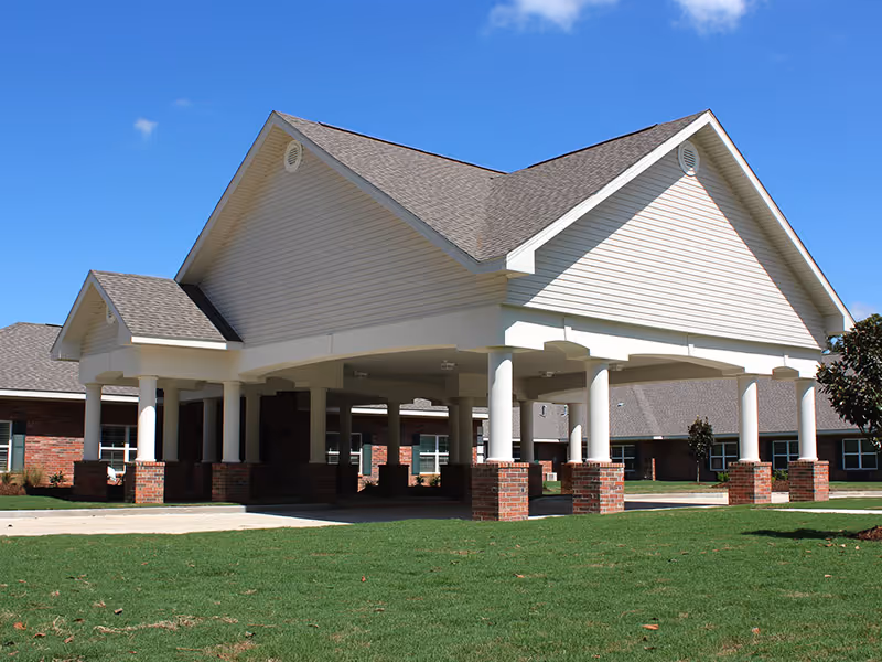 Exterior view of a senior living facility entrance with a large covered drop-off area supported by white columns on brick bases, surrounded by green grass and clear blue sky.