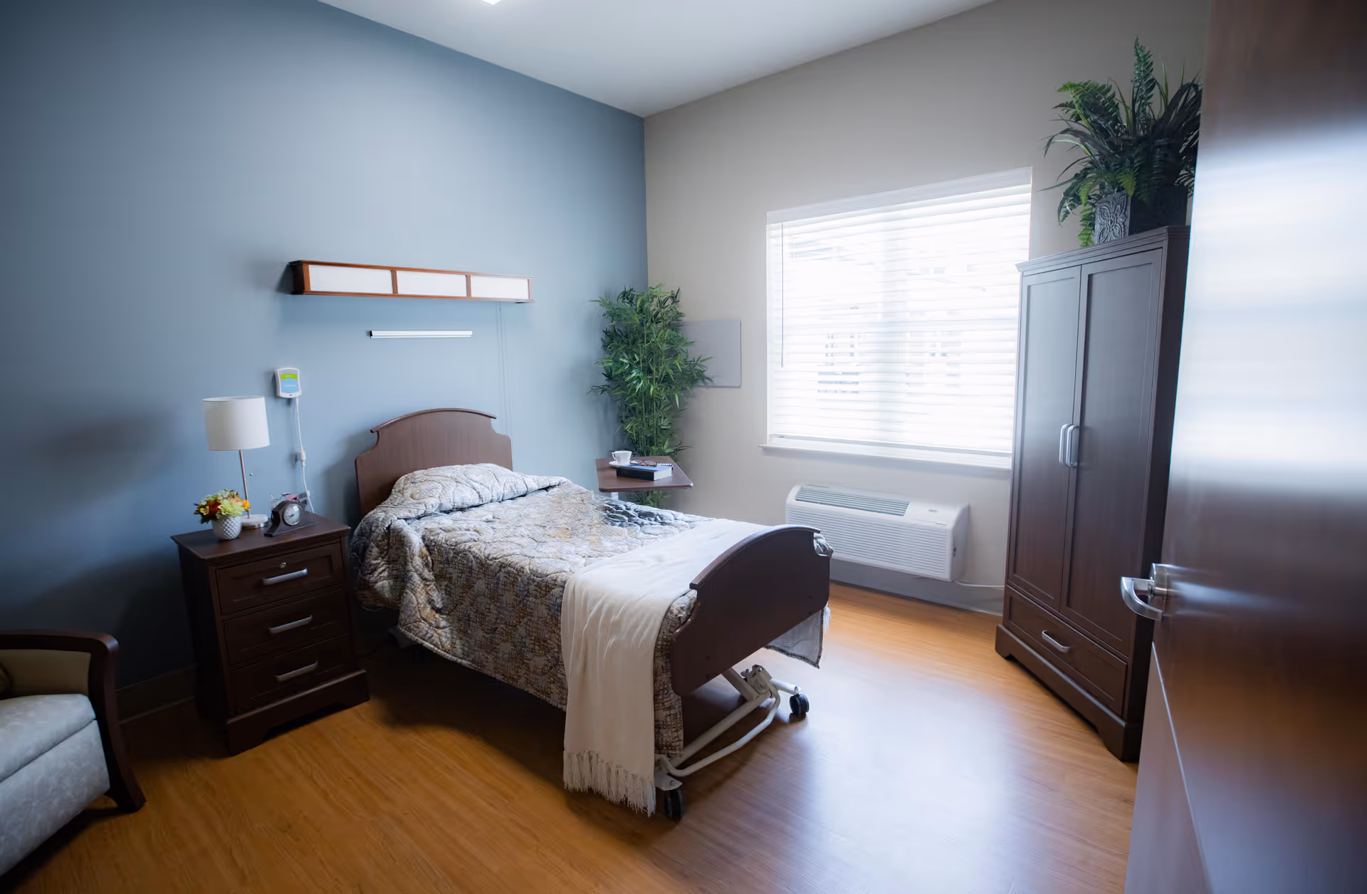 A neatly arranged bedroom in a senior living facility with a single bed covered in a patterned quilt, a wooden nightstand with a lamp and clock, a large window with blinds, a wooden wardrobe, a potted plant, and a comfortable chair.