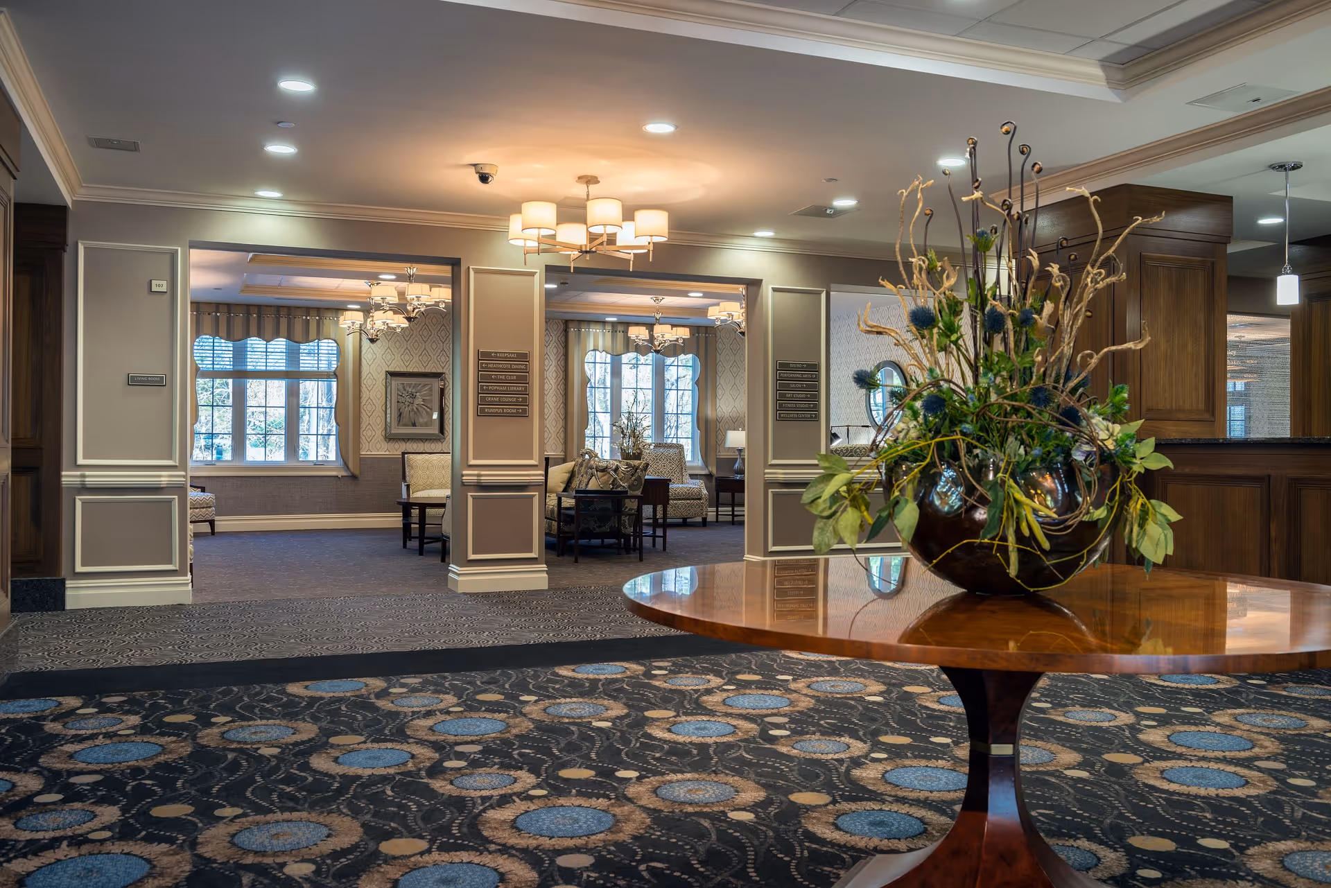 Interior view of a senior living facility lobby area with a round wooden table in the foreground holding a large decorative floral arrangement. The background shows an open seating area with armchairs, tables, large windows with curtains, and soft ceiling lighting fixtures. The walls are decorated with molding and signage indicating directions to various rooms.
