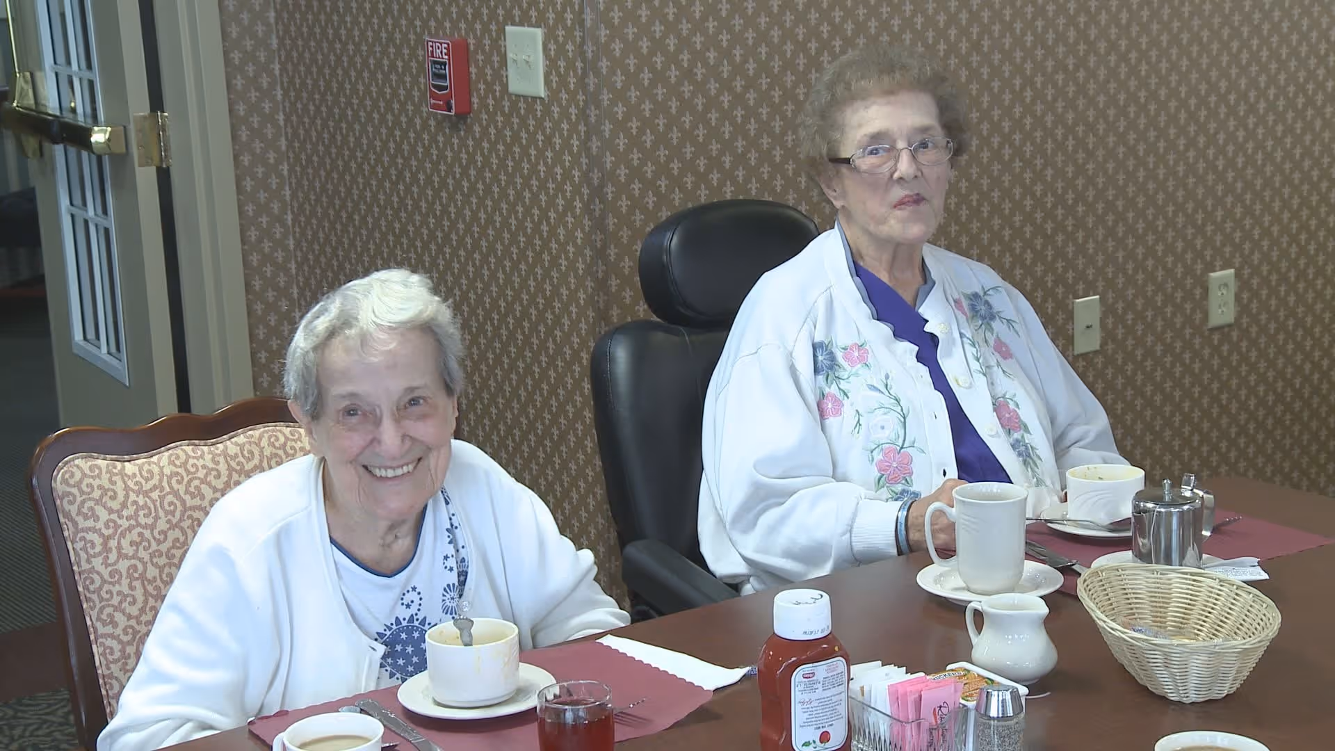 Two elderly women sitting at a dining table in a senior living facility. One woman is smiling and wearing a white cardigan, while the other woman is wearing glasses and a white jacket with floral embroidery. The table has cups, a glass of juice, a basket, and condiments on it. The background shows patterned wallpaper and a fire alarm on the wall.