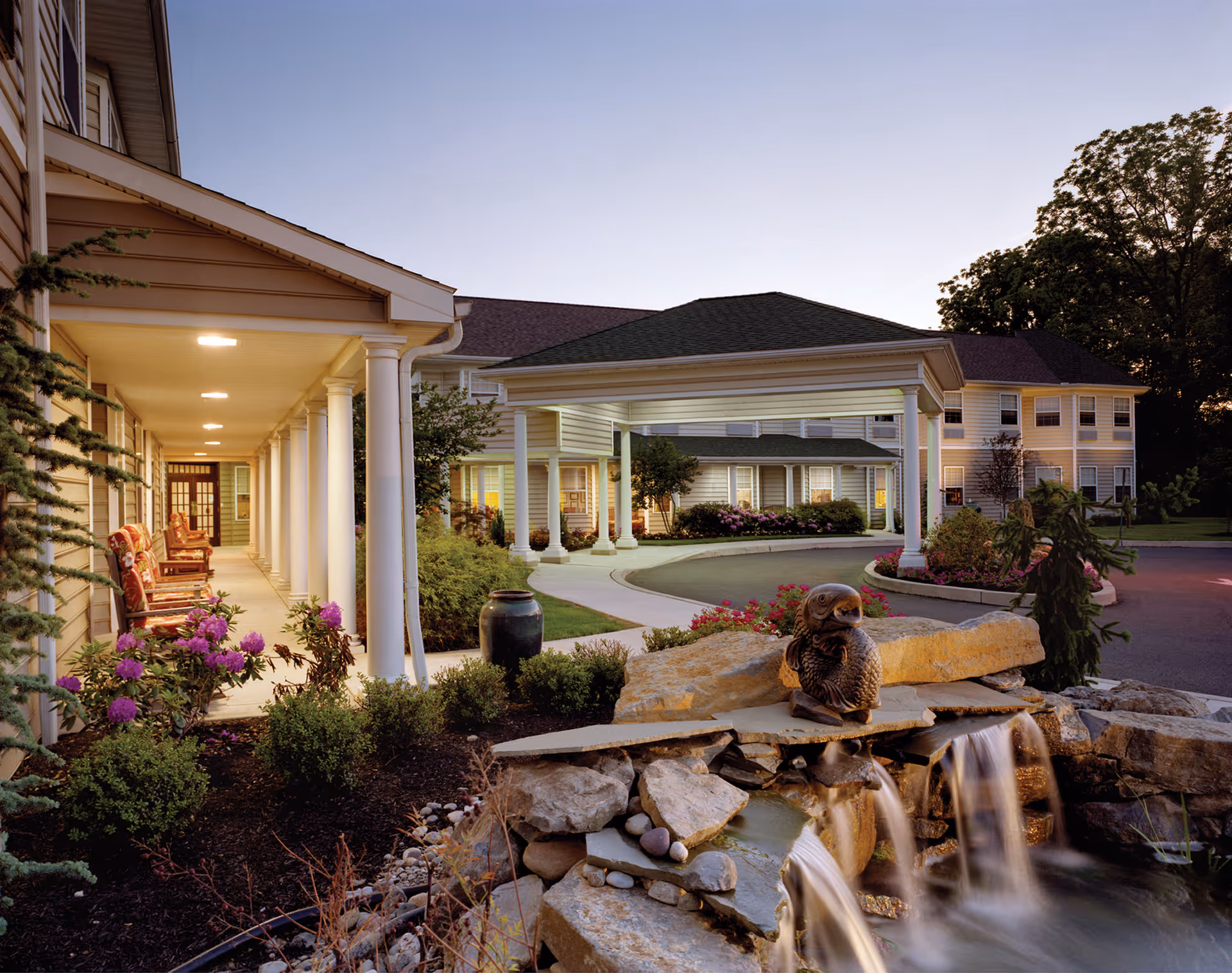 Exterior view of Dresher Estates by Priority Life Care at dusk, showing a covered entrance with white columns, a landscaped garden with flowers and shrubs, a small waterfall feature with a bird sculpture, and a multi-story building in the background with lit windows.