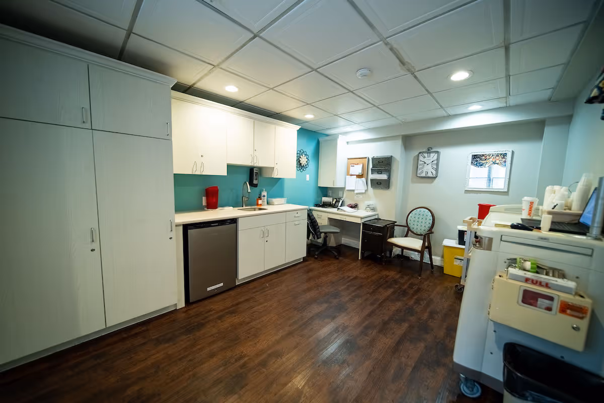 Interior view of a room with white cabinets, a countertop with a sink, a dishwasher, and a small desk area with a chair. The room has wood flooring, a clock on the wall, a window with a floral valance, and various office and medical supplies on a cart and desk.