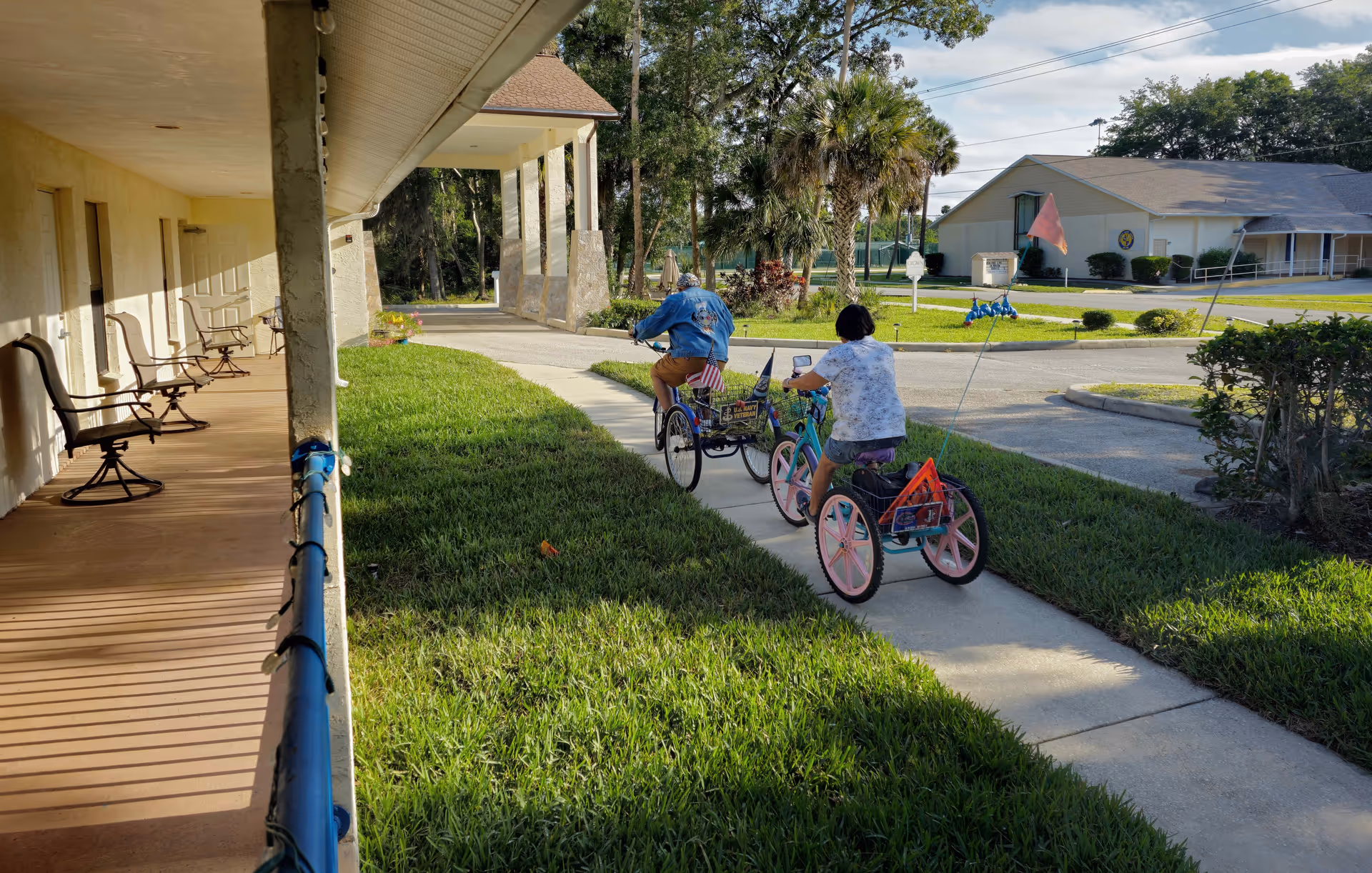 Two people riding tricycles on a sidewalk next to a building with a covered porch and chairs. The area is grassy with trees and another building visible in the background under a partly cloudy sky.