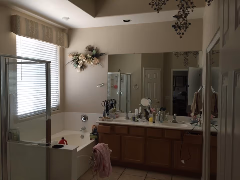 A bathroom with a large mirror above a double sink vanity with wooden cabinets. There is a bathtub with a shower enclosure next to a window with blinds and a valance. Various toiletries and personal items are on the countertop. A floral wall decoration is above the bathtub, and towels hang on the wall.