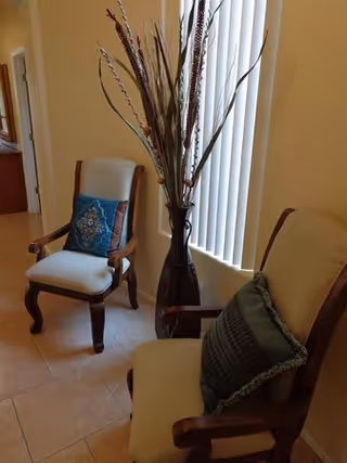Two wooden armchairs with cushions placed near a window with vertical blinds. Between the chairs is a tall decorative vase with dried branches. The room has beige walls and tiled flooring.