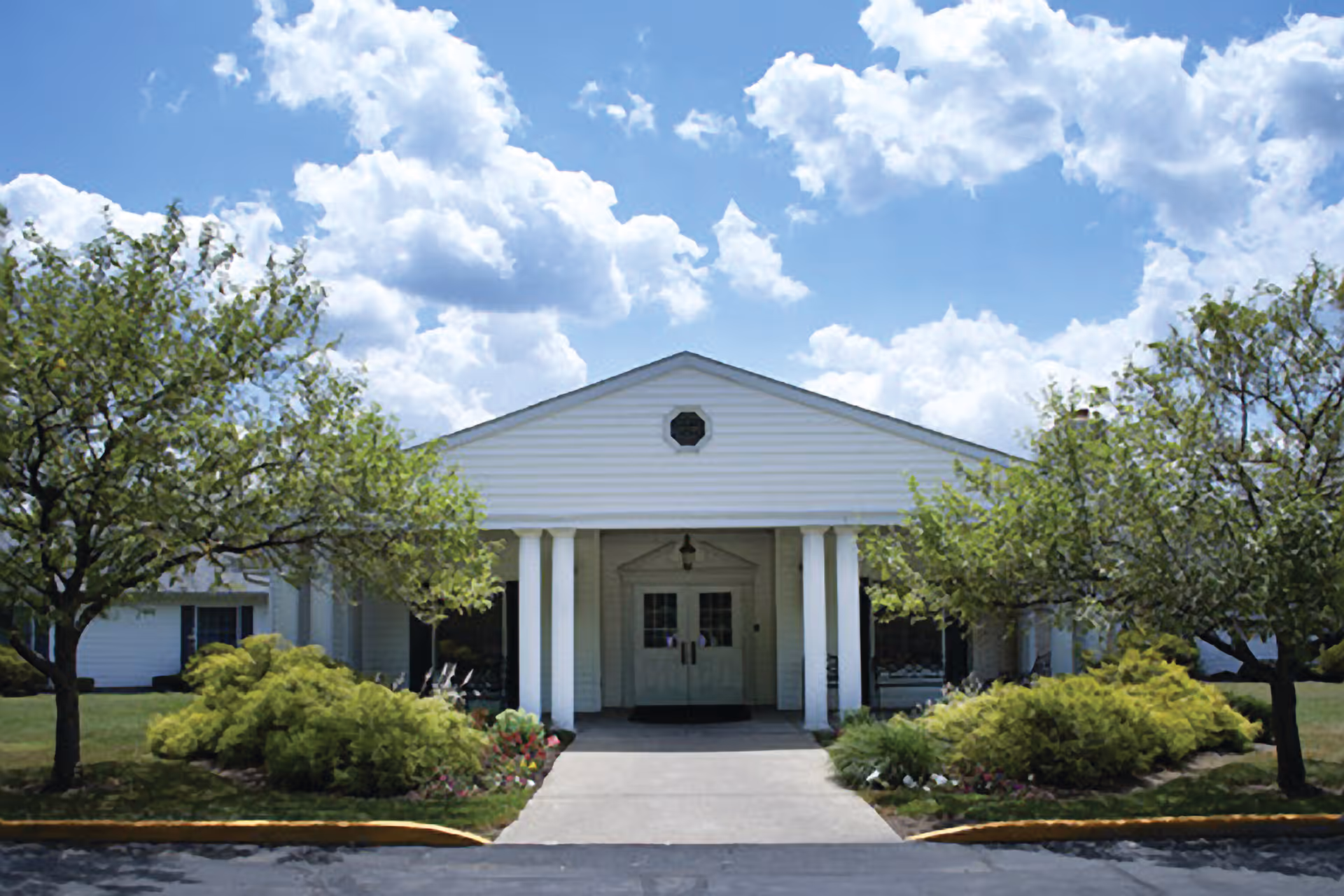 Front exterior view of a single-story white building with a triangular pediment supported by four white columns. There are two trees and landscaped bushes with flowers on either side of the concrete walkway leading to double entrance doors. The sky is blue with scattered clouds.