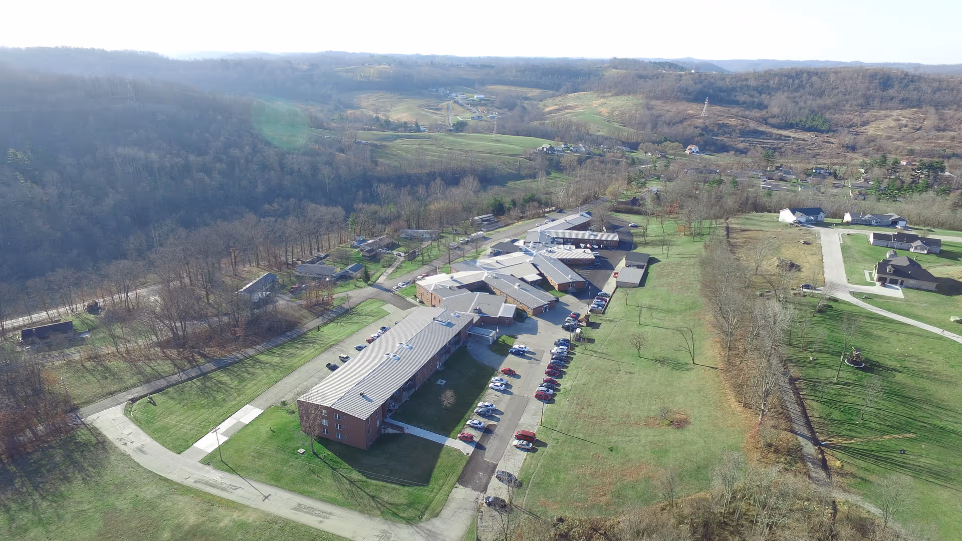 Aerial view of Altercare Summit Acres facility surrounded by green lawns and trees, with multiple connected buildings and a parking lot with several cars. The landscape includes rolling hills and scattered houses in the background under a clear sky.