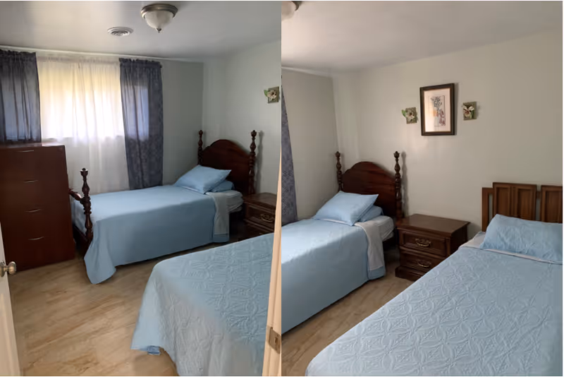A small bedroom with two single beds covered in light blue bedspreads. One bed has a wooden headboard with turned posts, and the other has a simpler wooden headboard. Between the beds is a wooden nightstand with two drawers. A framed picture and two small decorative wall hangings are above the nightstand. A wooden dresser is positioned near a window with sheer white curtains and darker blue drapes. The floor is light-colored wood or laminate.