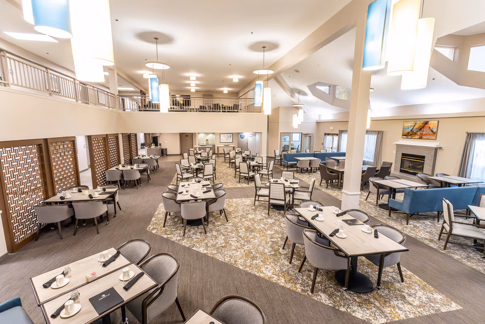 Spacious dining room in Asher Point Independent Living of Tyler featuring multiple tables with chairs, set with cups and napkins. The room has high ceilings with modern hanging lights, a fireplace, large windows with curtains, and a mezzanine level overlooking the area.