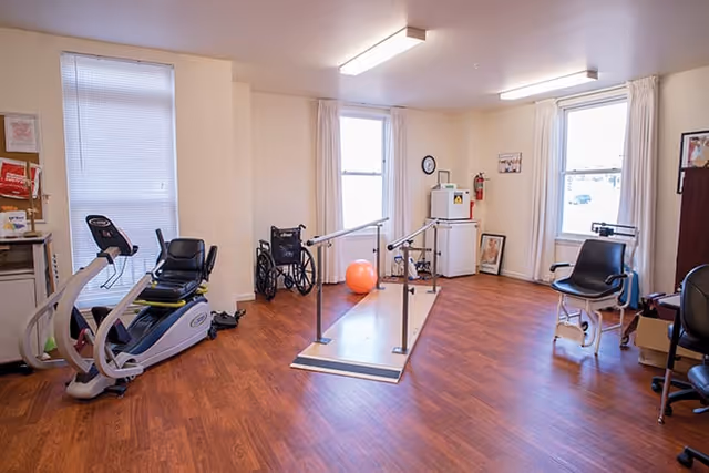 A physical therapy or rehabilitation room with wooden flooring, featuring exercise equipment including a recumbent bike, parallel bars for walking practice, a wheelchair, an exercise ball, and a chair with leg weights. The room has three windows with white curtains and light beige walls.