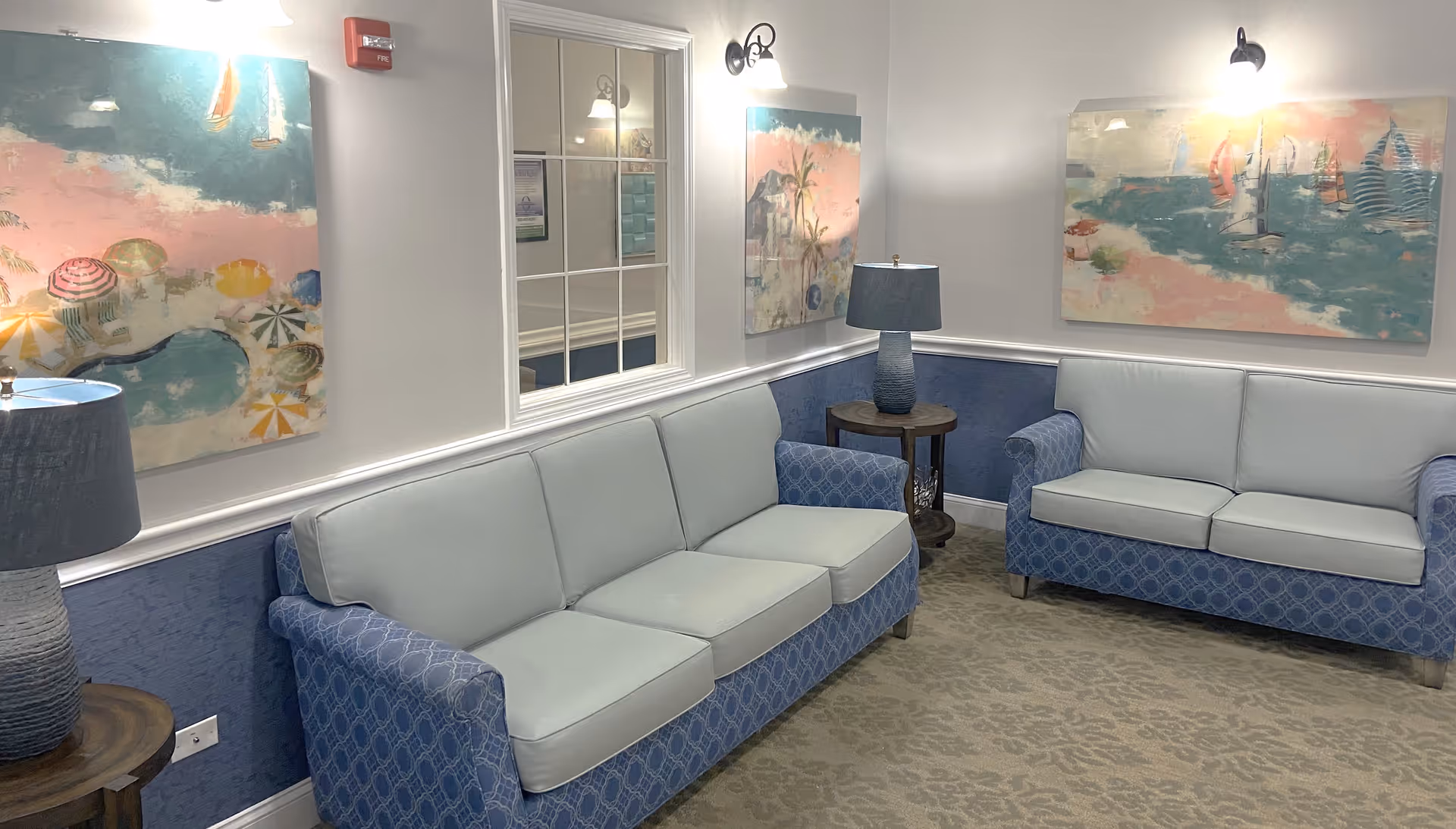 A cozy sitting area with two blue patterned sofas with light blue cushions, two side tables each with a blue lamp, and three colorful paintings on the walls depicting beach and ocean scenes. The walls are painted white with a blue wainscoting, and there is a window with white framing between the paintings.