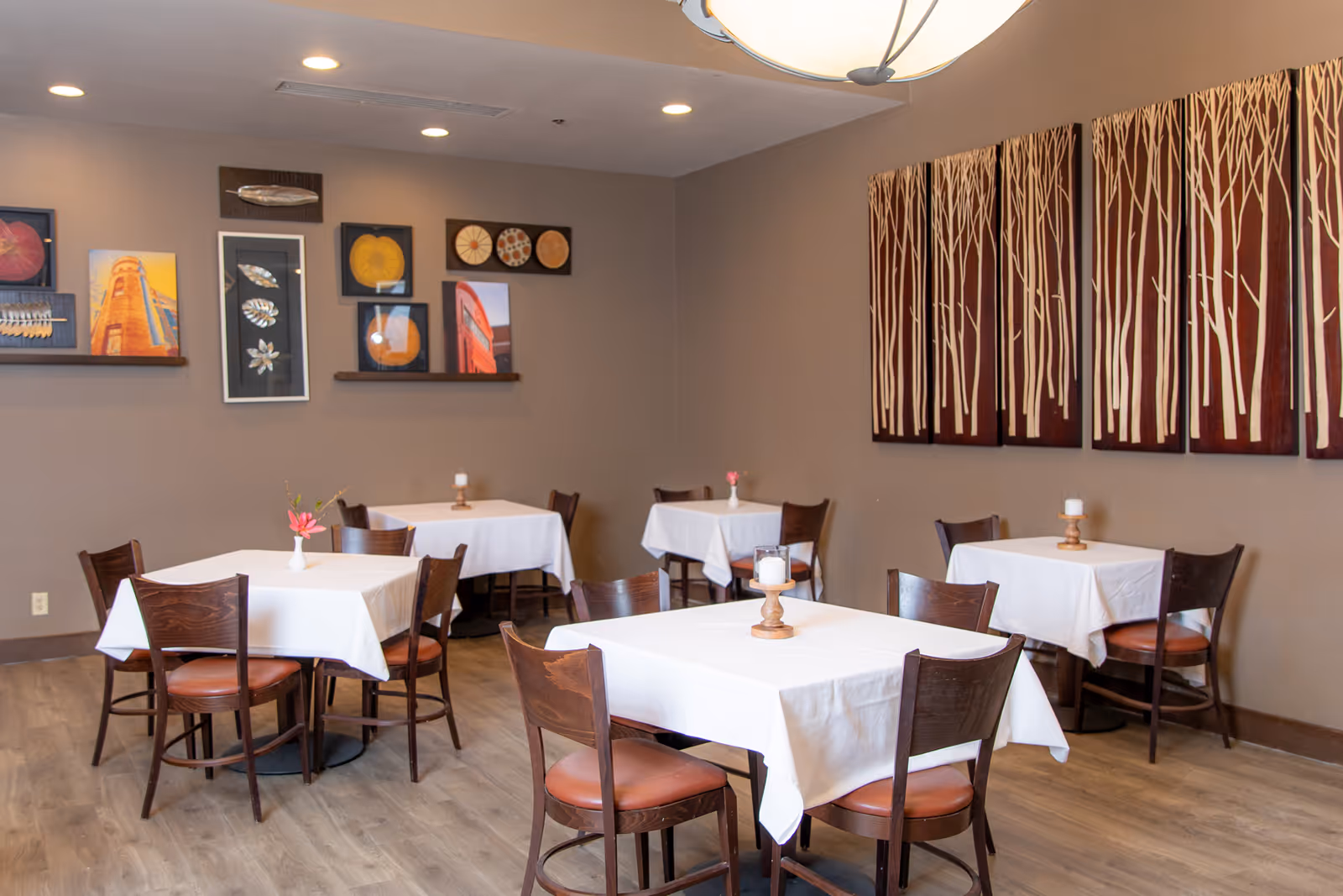 A dining room with several square tables covered with white tablecloths, each surrounded by four wooden chairs with brown seats. The walls are decorated with framed artwork and a large wooden panel featuring a tree design. The floor is wooden, and the room is softly lit by ceiling lights.