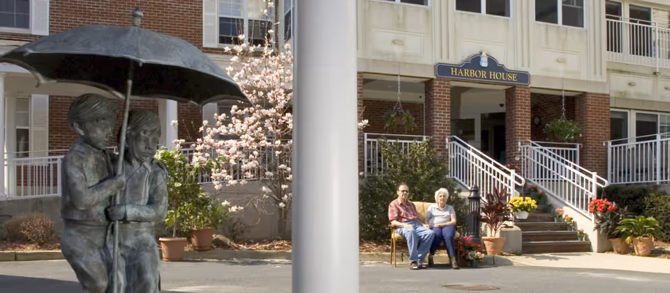 Two elderly people sitting on a bench outside the entrance of Harbor House Assisted Living, with a statue of two children holding an umbrella in the foreground and flowering plants around the entrance.