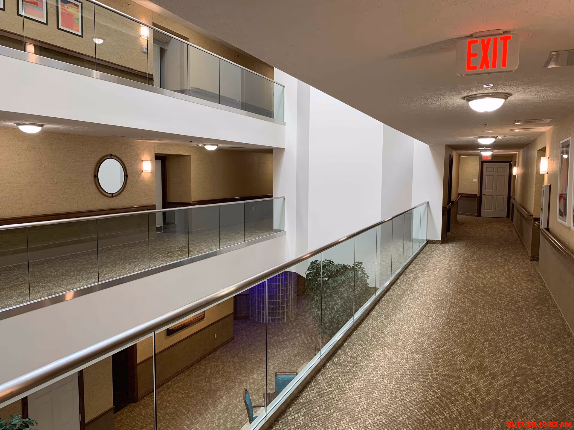 Interior hallway of Aspen Trail Retirement Resort with beige carpet and walls, glass railings overlooking a lower floor, ceiling lights, framed pictures on the walls, and an illuminated red EXIT sign at the end of the corridor.