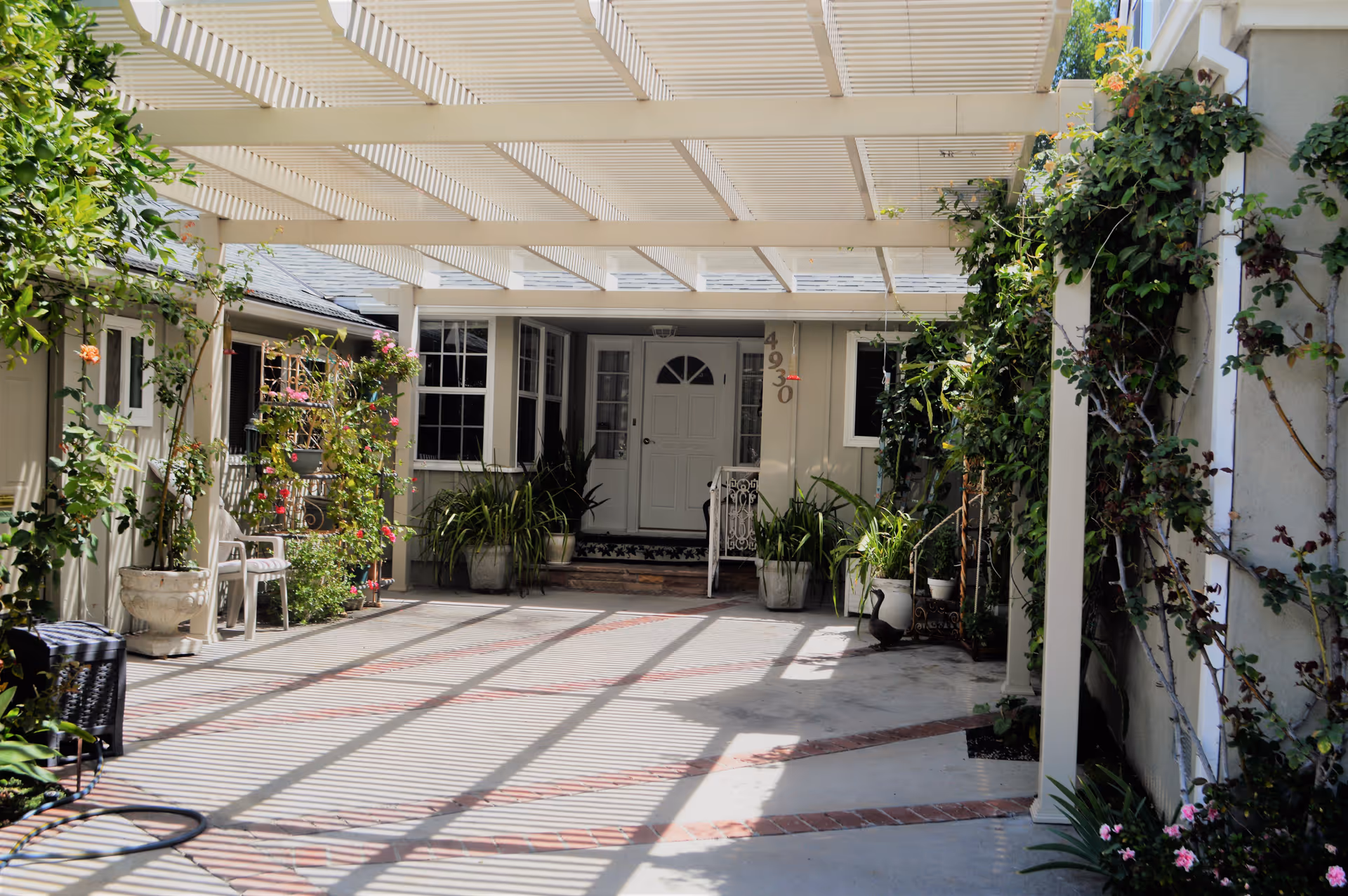 A covered outdoor patio area with a white pergola casting shadows on the ground. The space is decorated with various potted plants and climbing vines along the walls. At the far end, there is a white door with windows on either side and the number 4930 displayed vertically next to the door.