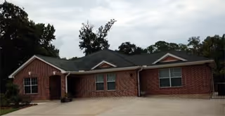 Single-story brick building with a dark shingled roof, multiple windows, and a driveway in front, surrounded by trees under a cloudy sky.