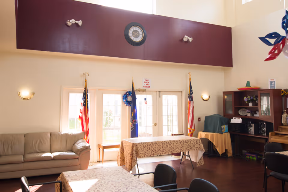 A bright common area in Whitehall Manor with a beige couch on the left, two tables covered with patterned tablecloths, and several chairs. There are two American flags and one state flag near glass doors letting in natural light. A clock is mounted on a maroon wall section above the doors, and there is a wooden cabinet with decorations and a covered item on the right side.