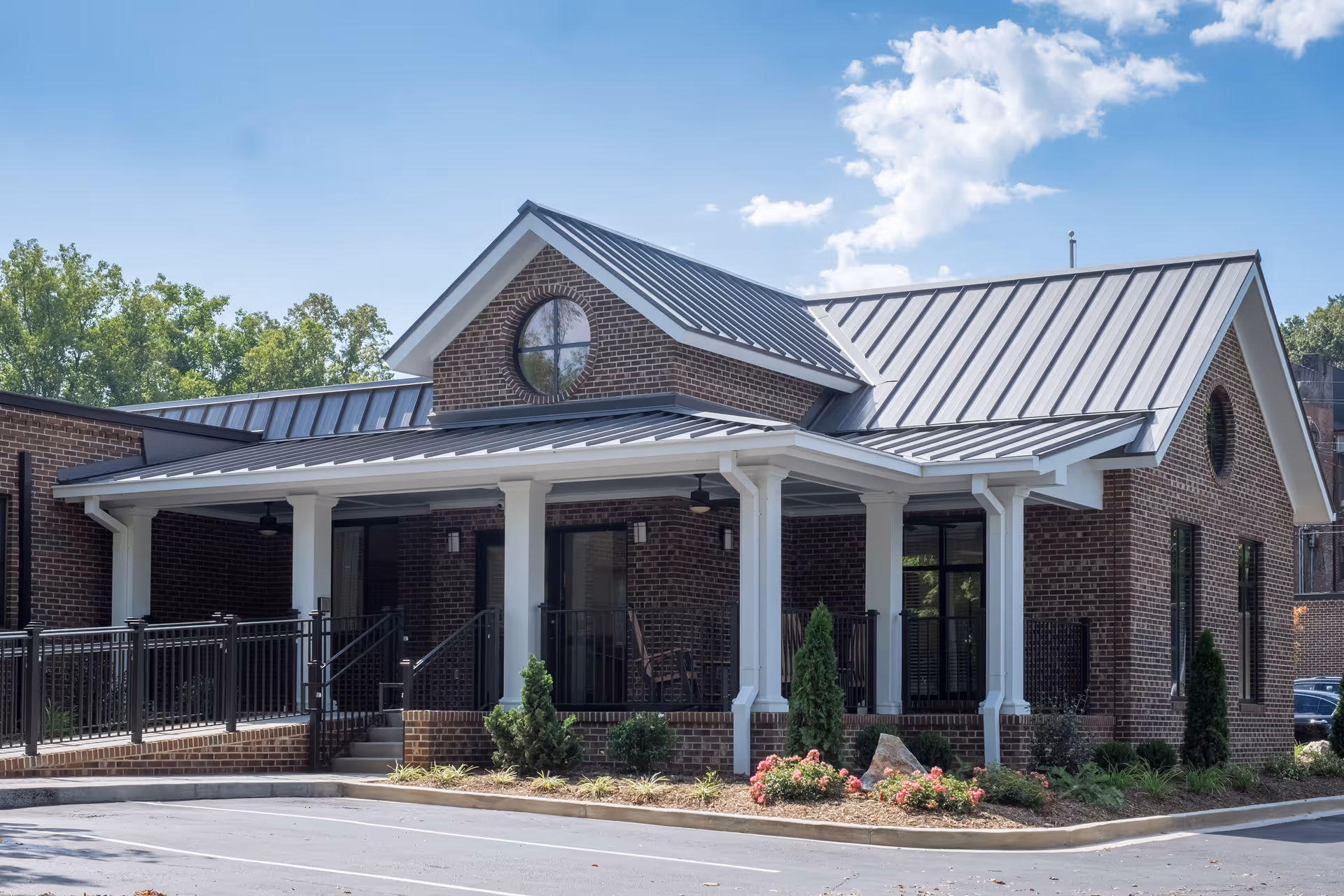 Exterior view of a brick building with a metal roof and white columns supporting a covered porch. The building has a circular window near the roof peak and is surrounded by landscaping with bushes and flowers. There is a paved parking area in front.