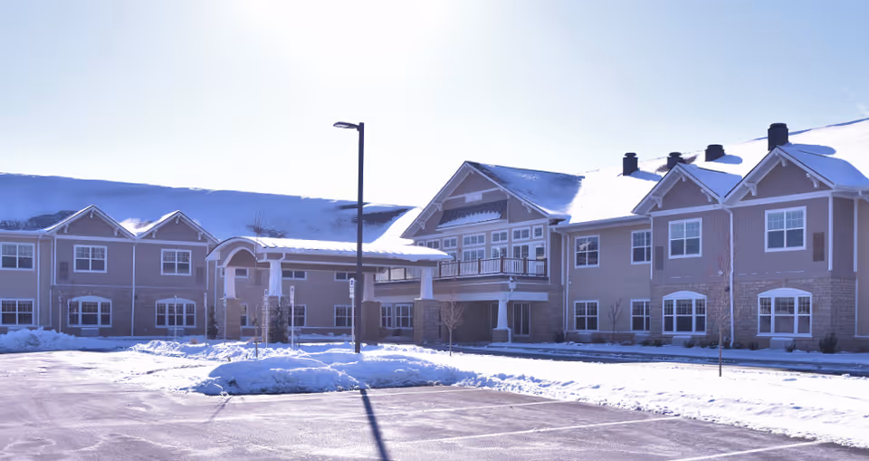 Exterior view of a senior living facility named Wooded Glen covered in snow with a clear sky. The building has multiple windows, a covered entrance, and a parking lot in front.