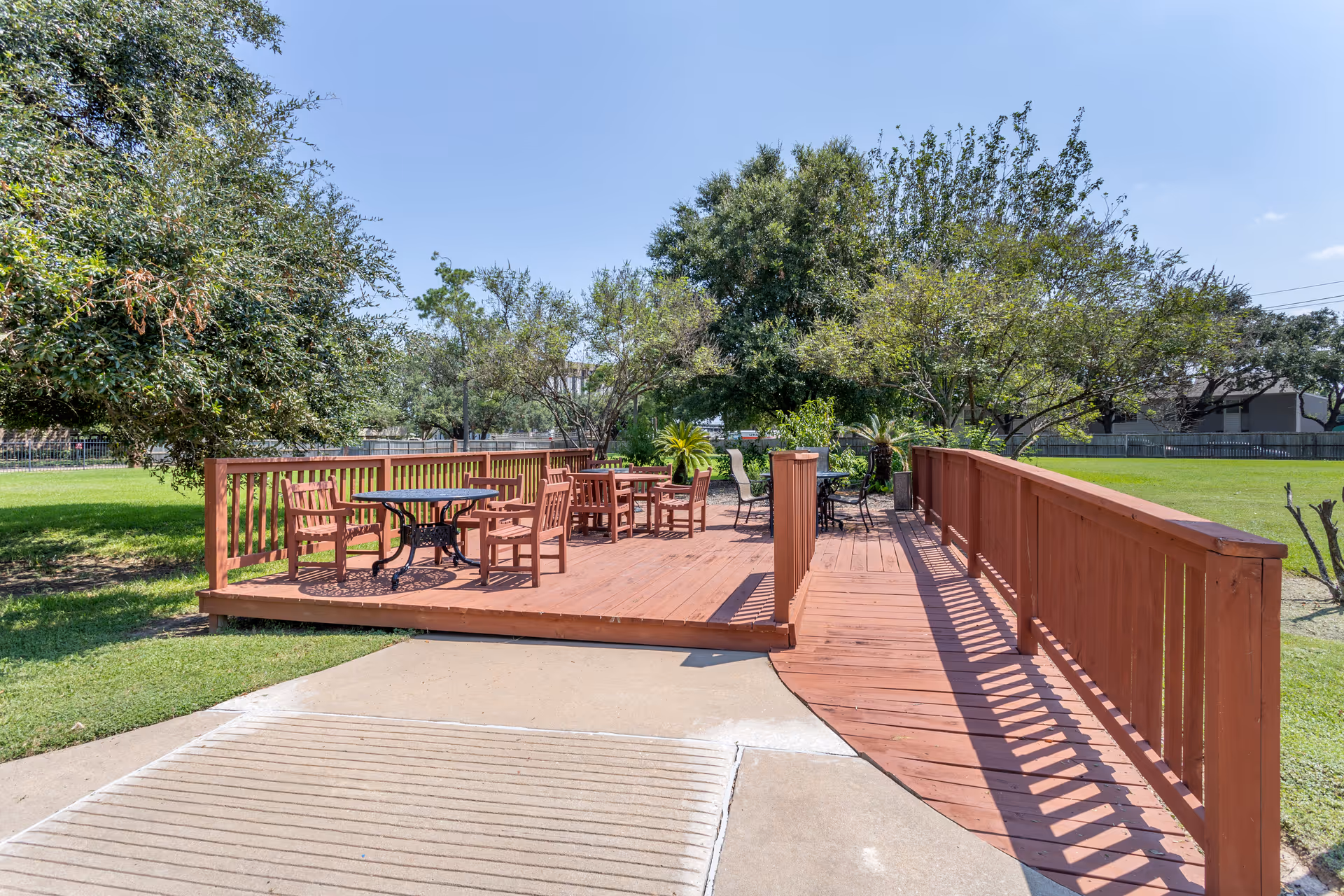 Outdoor wooden deck area with several tables and chairs surrounded by green grass and trees under a clear blue sky.