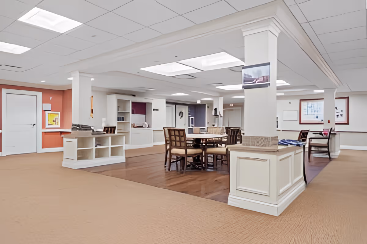 A spacious interior common area in a senior living facility with a round wooden table surrounded by chairs in the center. The room features white pillars, built-in shelving, a mix of carpet and wood flooring, and overhead fluorescent lighting. There are framed pictures on the walls and a small TV mounted on one of the pillars.
