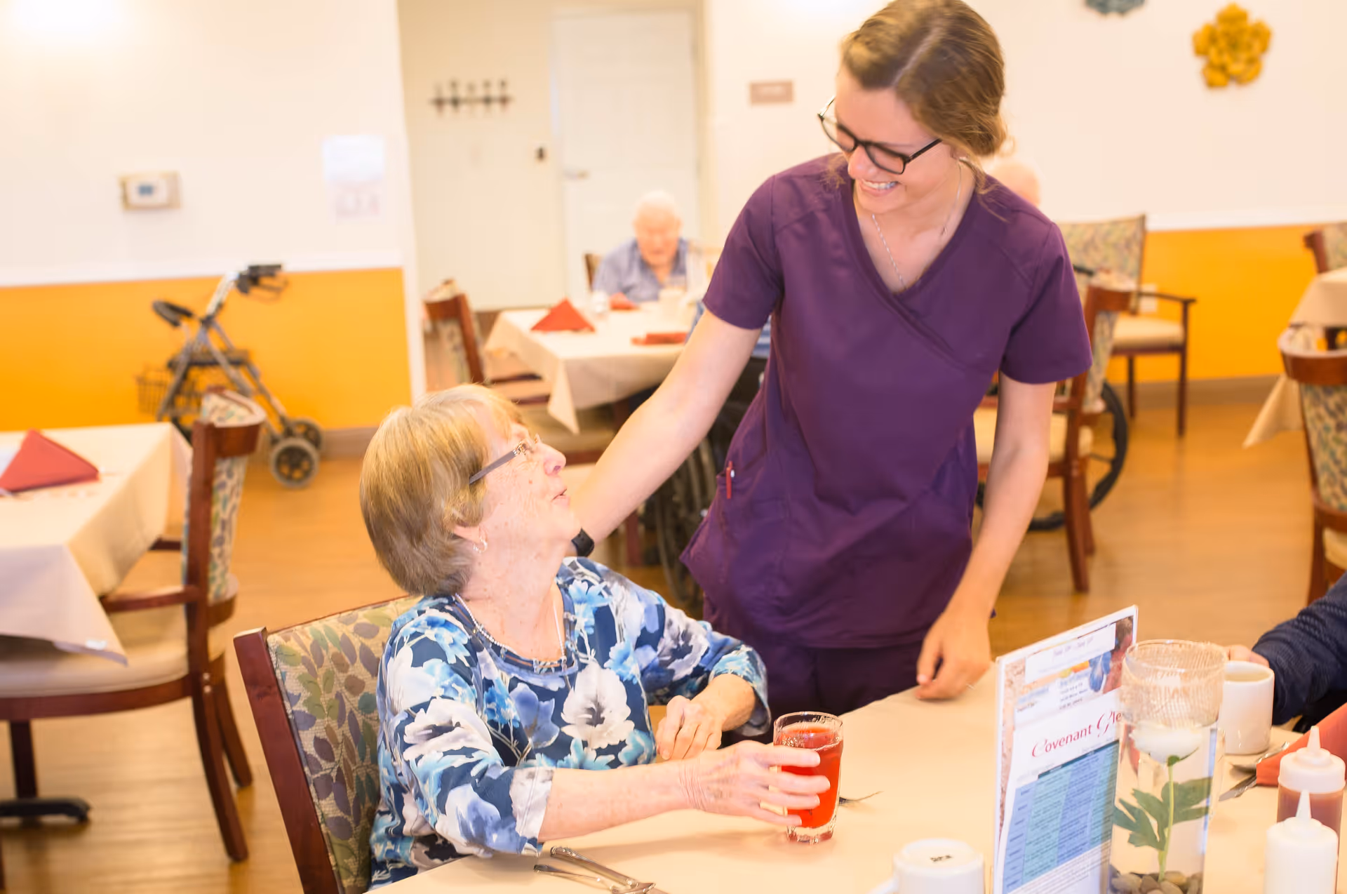 A smiling caregiver in purple scrubs interacts warmly with an elderly woman seated at a dining table holding a glass of red drink in a senior living facility dining room. The room has tables with white tablecloths and red napkins, and another elderly person is visible in the background.