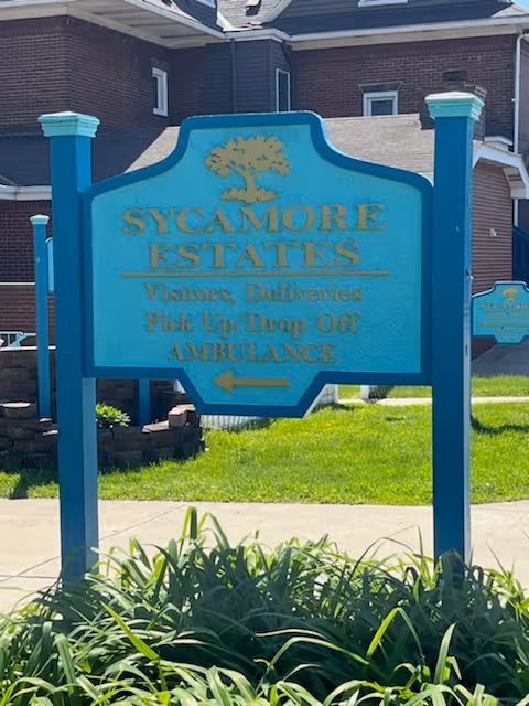 A blue and teal sign with gold lettering in front of a brick building. The sign reads 'SYCAMORE ESTATES Visitors, Deliveries Pick Up/Drop Off AMBULANCE' with an arrow pointing left. There is greenery and plants in front of the sign and a sidewalk behind it.