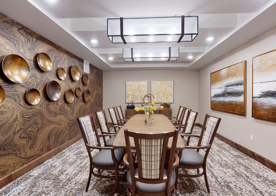 A dining room with a long wooden table surrounded by ten chairs with patterned upholstery. The room features a decorative wall with multiple bronze circular plates on the left, two abstract paintings on the right wall, and two modern light fixtures on the ceiling. A vase with yellow flowers is placed in the center of the table.